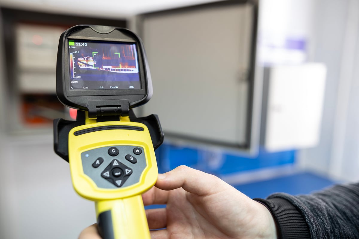 Electrician holding a thermal imaging camera to inspect an open electrical panel for overheating components during preventive maintenance